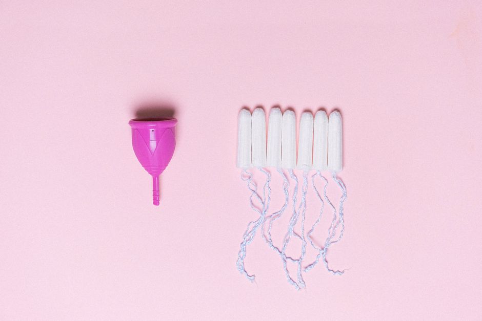 A pink menstrual cup situated on the left side of the image, with its stem pointing downward, placed on a soft pink background. To the right, there is a group of six white sanitary pads arranged in a 