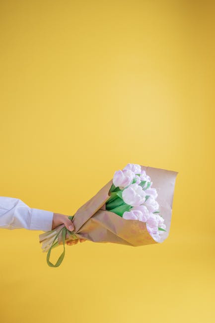 A person in a white shirt is holding a bouquet of fresh pink tulips wrapped in light brown kraft paper and secured with a green ribbon. The bouquet consists of multiple tulip stems with delicate, slig