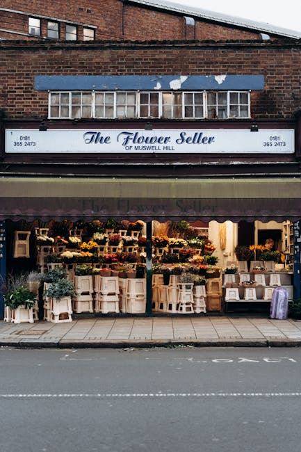 The storefront of The Flower Seller of Muswell Hill features a large display of fresh floral arrangements arranged on open shelves and white stools outside the shop. The flowers include vibrant yellow