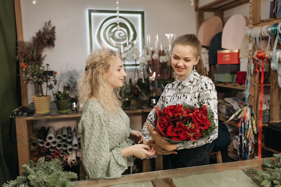A young woman with light brown hair tied back smiling as she receives a floral gift, a bouquet of red roses, from another woman with curly blonde hair inside a florist shop. The bouquet is wrapped wit