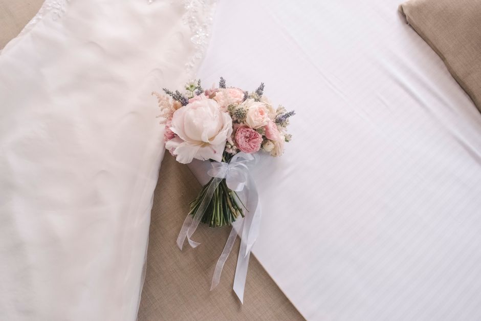 A delicate floral arrangement featuring soft pink and white peonies, roses, and lavender sprigs, arranged in a rounded bouquet with visible green stems bound by a translucent white ribbon. The bouquet