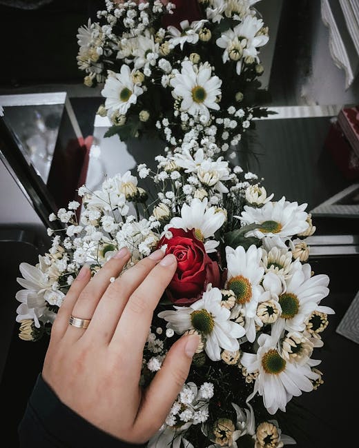 A floral arrangement featuring a bouquet of white daisies with yellow centers, baby's breath, and a single red rose, held by a hand wearing a ring, positioned on a reflective glass surface. The bouque