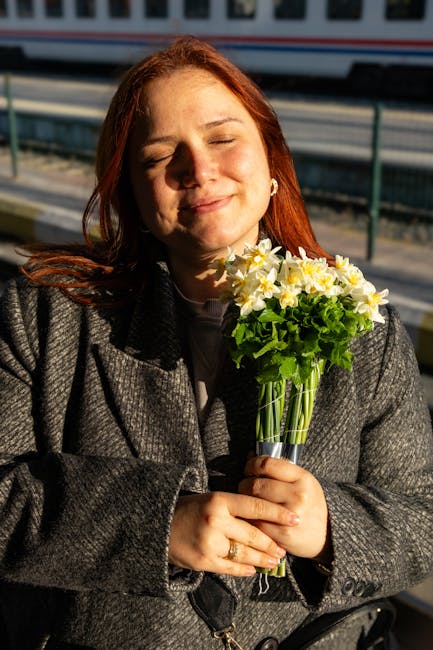 A woman with red hair, softly smiling with her eyes closed, holds a vibrant bouquet of fresh yellow daffodils and green foliage in her hands. The flowers are arranged in a clear glass vase with a silv
