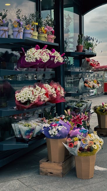 A display of various fresh floral arrangements at a flower shop, featuring bouquets wrapped in colorful paper and placed on dark metal shelving units. The arrangements include white chrysanthemums, pi