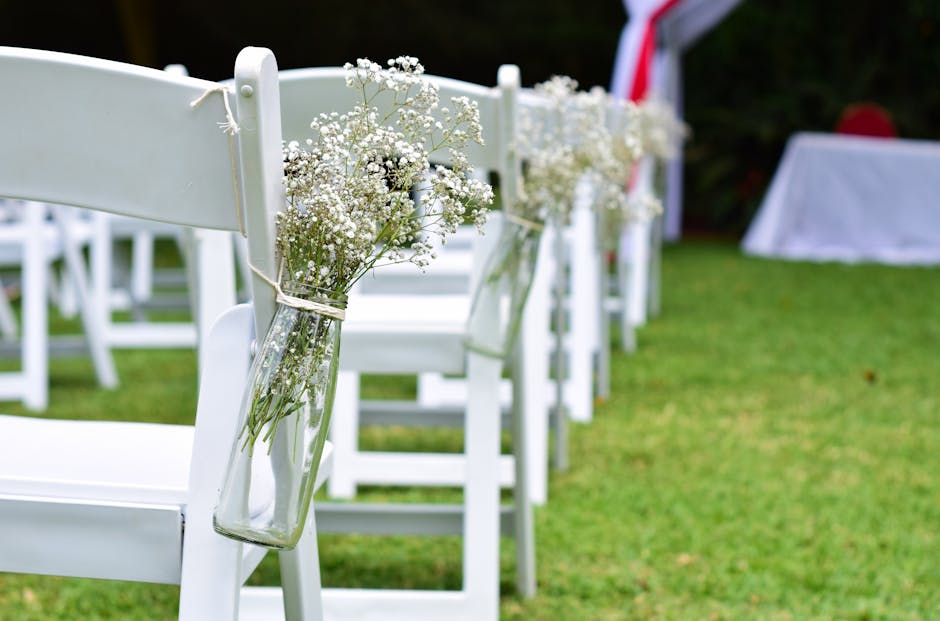 A row of white wooden chairs arranged outdoors on a grassy area, each decorated with a clear glass jar tied to the backrest containing small bouquets of delicate white baby's breath flowers. The jars