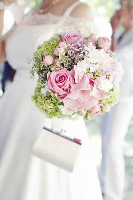 A close-up of a bridal bouquet featuring soft pink roses, white hydrangeas, and pale pink spray roses, accented with delicate white baby's breath and variegated green leaves. The bouquet is bound with