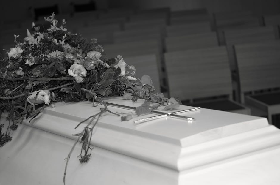 A white coffin adorned with a bouquet of fresh flowers, including roses and greenery, rests on top during a funeral service. The flowers are arranged in a loose, natural style with some petals and lea