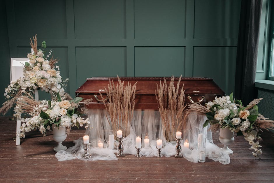 A floral arrangement featuring white and cream roses, hydrangeas, and greenery, complemented by tall, pale beige pampas grass, all set in elegant white vases on either side of a closed wooden casket.